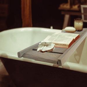 Puede incluir: Una bañera blanca con una bandeja de madera en la parte superior. La bandeja tiene un libro, una esponja blanca y un vaso de agua.
