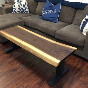 May include: A long, live edge walnut wood coffee table with a natural wood finish and black metal legs. The table is sitting in front of a brown sectional sofa with pillows.