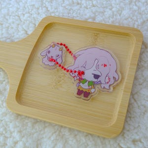 May include: A clear acrylic keychain featuring a cartoon character with pink hair and a small goat. The keychain is connected by a red beaded chain. The character is wearing a green and yellow outfit. The keychain is on a light brown wooden tray.
