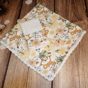 May include: A white baby blanket with a crocheted border. The blanket features a floral pattern with yellow, orange, and green flowers and a rocking horse design.