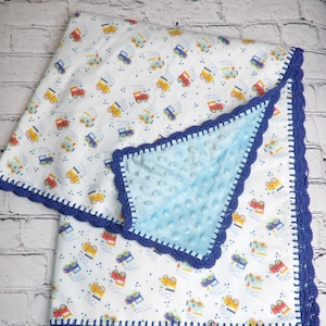 May include: A baby blanket with a blue and white minky fabric on one side and a white fabric with a repeating pattern of toy trucks on the other side. The blanket is edged with a blue crocheted border.