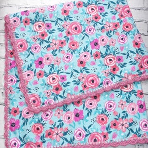 May include: A floral baby blanket with a light blue background and a pattern of pink and fuchsia roses and other flowers. The blanket has a pink crocheted border.