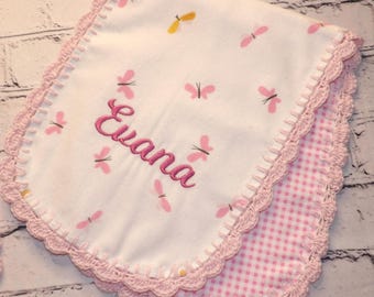 May include: A white baby blanket with pink butterflies and the name "Evana" embroidered in pink. The blanket has a pink gingham backing and a pink crocheted border.