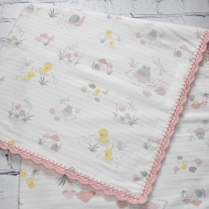 May include: A white baby blanket with a pink crocheted border. The blanket features a repeating pattern of ducks, frogs, and snails on a light pink and white striped background.