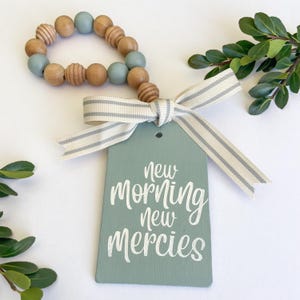 May include: A light green wooden tag with the text "new morning new mercies" in white paint. The tag is tied with a white and gray striped ribbon and a beaded garland of light brown and light blue beads.