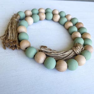 May include: A decorative wooden bead garland with alternating sage green and natural wood beads. The garland is accented with a jute tassel and jute rope. The beads are approximately 1 inch in diameter.