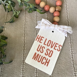 May include: White wooden sign with the text "He loves us so much" in orange lettering. The sign is tied with a white and brown gingham ribbon and is hanging from a strand of wooden beads.