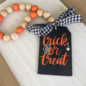 May include: A black wooden sign with the text "trick or treat" in orange lettering. A white spider and spider web are also on the sign. The sign is attached to a black and white checkered ribbon and a wooden bead garland with orange and natural wood beads.