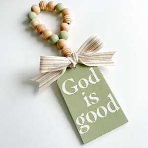 May include: A decorative wooden tag with the text "God is good" in white lettering. The tag is sage green and attached to a beaded garland with a striped ribbon bow. The beads are a mix of natural wood and sage green.