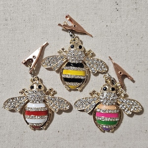 Puede incluir: Tres clips decorativos con forma de abeja con alas y cuerpos de metal dorado. Una abeja tiene un cuerpo a rayas amarillas y negras, otra tiene rayas rojas y blancas, y la tercera tiene rayas rosas, verdes y moradas. Cada clip tiene un pequeño cierre de metal.