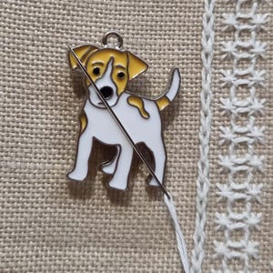 Könnte beinhalten: Ein weißer und gelber Email-Hundeanhänger mit einer Nadel und einem Faden, die durch ihn gehen. Der Anhänger befindet sich auf einem beigefarbenen Stoffhintergrund.