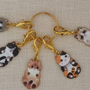 May include: Six cat-shaped charms with gold-coloured clasps and a gold ring. The charms feature various cat designs in colours like grey, black, orange, and brown. The charms are likely for jewellery or crafting.