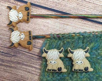 May include: Highland cow-themed knitting needle toppers and stitch markers. The toppers are brown with white fur and black accents. The stitch markers are tan with black accents. The needles are multicolored.
