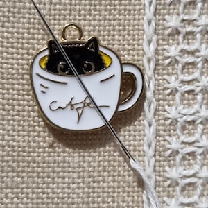 May include: A white enamel charm in the shape of a coffee cup with a black cat peeking out. The cup has gold trim and the word "caffe" is written on the front. A needle is threaded with white thread, next to a cross-stitch pattern.