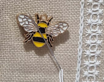 May include: A gold bee charm with black and yellow stripes and white wings is being sewn onto a beige fabric with a white crocheted border.