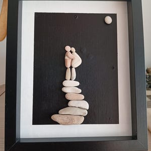 May include: Framed artwork featuring a black background with a white border. The artwork depicts a couple embracing, crafted from smooth, light-colored stones. A stack of stones forms a base, and a small, round stone represents the moon.