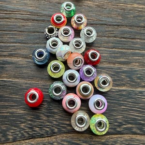 May include: A collection of colorful glass beads with silver-toned metal cores. The beads are various colors, including red, green, pink, and purple, and are scattered on a dark wooden surface. These beads are suitable for jewelry making.
