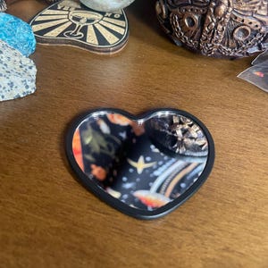 Può includere: Uno specchio a forma di cuore con una cornice nera. La superficie dello specchio riflette un colorato motivo floreale e celeste. Lo specchio è su una superficie di legno, con altri oggetti decorativi sullo sfondo.