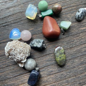 Puede incluir: Una colección de varias piedras preciosas y cristales, incluyendo una esfera de cuarzo rosa, un colgante de arenisca azul y una piedra de jaspe rojo. Las piedras son de diferentes formas y tamaños, con herrajes de metal plateado.