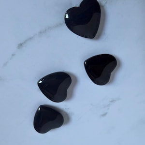 May include: Three polished black heart-shaped stones with a smooth, reflective surface. Each stone features a rounded, symmetrical heart silhouette with a deep, solid black color and a glossy finish that catches the light across its curved edges.