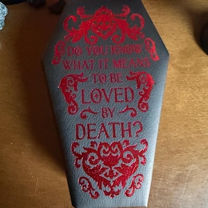 Puede incluir: Una caja gris en forma de ataúd con detalles decorativos rojos y el texto "DO YOU KNOW WHAT IT MEANS TO BE LOVED BY DEATH?" La caja tiene una superficie texturizada y probablemente se utiliza para almacenamiento o exhibición.