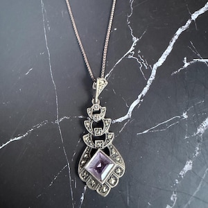 May include: A silver-toned pendant necklace featuring a square-cut light purple gemstone at the base. The pendant has a tiered, geometric design embellished with small, dark, faceted stones set in metal, suspended from a thin silver-toned chain.