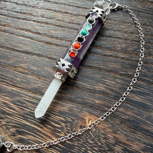 May include: A purple crystal pendulum with a silver chain. The pendulum features seven colored stones, representing the chakras, set in a silver metal casing. The crystal point is clear and faceted.
