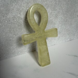 May include: A pale yellow, translucent ankh symbol carved from stone. The object features a smooth, polished surface with a rounded loop at the top and a vertical cross shape extending downward, displaying natural crystalline inclusions throughout.