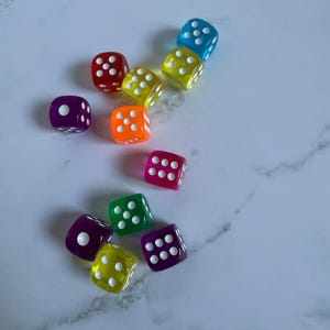 May include: A collection of eleven translucent, six-sided dice in various colors including red, yellow, blue, orange, purple, pink, and green. Each die features rounded corners and white pips representing numbers one through six on their faces.