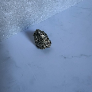 May include: A raw, metallic pyrite mineral specimen with a cubic, crystalline structure and a reflective, brassy gold surface. The cluster features sharp, angular edges and a dense, textured appearance with natural variations in light reflection.