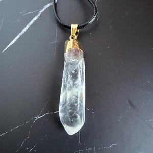 May include: A clear, elongated, raw crystal pendant suspended from a thin black cord. The crystal features a natural, irregular shape with visible internal inclusions and is capped with a polished gold-toned metal bail at the top.