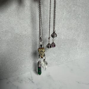 May include: Two silver-toned metal chains featuring multiple pendants. One chain holds a triangular dalmatian jasper stone, a green and magenta zoisite point, and a clear heart-shaped charm. The other chain features two silver claw-style settings.