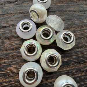 Puede incluir: Una colección de cuentas decorativas con detalles metálicos plateados. Las cuentas varían en forma y color, incluyendo blanco nacarado, iridiscente y transparente con purpurina. Para la fabricación de joyas o manualidades.