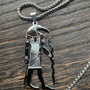 May include: A silver-toned pendant necklace featuring an Egyptian Thoth design. The pendant depicts the deity with a bird head, black body, and white accents. The necklace has a chain with a textured, rounded link design.