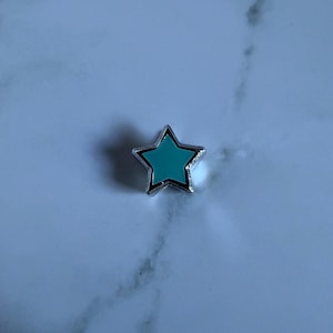 May include: A small, five-pointed star charm featuring a vibrant turquoise enamel center encased in a polished silver-tone metal border. The star has a clean, symmetrical shape with defined edges and a smooth, glossy finish on the front surface.