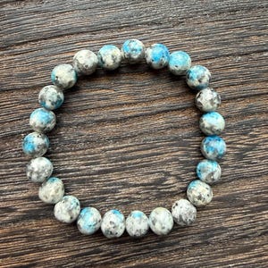 Puede incluir: Un brazalete de cuentas redondas, moteadas, en tonos blanco, gris y azul. Las cuentas tienen un aspecto veteado. El brazalete tiene forma circular. Las cuentas están ensartadas.