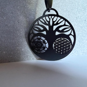 May include: A black circular pendant on a matching chain featuring a cutout tree silhouette. The design incorporates a geometric Metatron's cube pattern on the left and a flower of life pattern on the right, all rendered in a flat black finish.