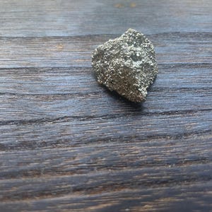 Op de afbeelding: Een klein, onregelmatig gevormd pyrietexemplaar met een metallic, goudachtige glans. Het ruwe oppervlak van het mineraal reflecteert licht, waardoor een sprankelend effect ontstaat. De pyriet ligt op een donkere, houten ondergrond.