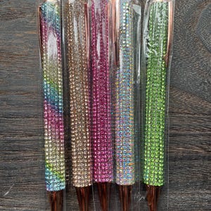 Könnte beinhalten: Sechs Stifte mit roségoldenen Akzenten und bunten Strasssteinen. Die Stifte sind einzeln in durchsichtigen Plastiktüten verpackt.