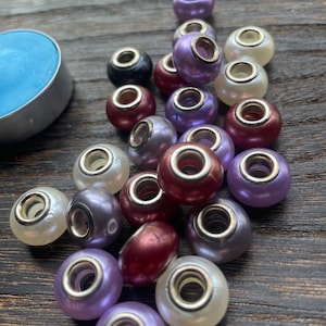May include: A collection of colorful beads in various shades of purple, red, black, and white. Each bead has a silver-colored metal core. The beads are scattered on a dark wooden surface, with a small blue candle in the background.