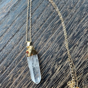 May include: A gold-colored chain necklace with a clear quartz crystal pendant. The crystal is a long, irregular shape with a gold-colored cap. The necklace is displayed on a dark wood surface.
