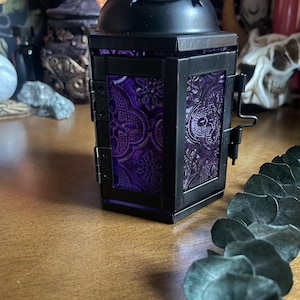 Puede incluir: Linterna de metal negro con una parte superior abovedada y un asa. La linterna presenta paneles de vidrieras moradas con un patrón ornamentado. La linterna está diseñada con una puerta con bisagras y un cierre metálico decorativo.