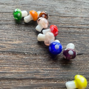 May include: A collection of colorful glass mushroom beads with white stems. The mushroom caps are various colors, including green, orange, brown, red, blue, purple, and yellow, each with white polka dots. The beads are arranged on a wooden surface.