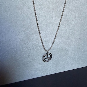 May include: A silver-toned ball chain necklace featuring a circular pendant. The pendant consists of a thin outer ring enclosing a complex, linear geometric symbol with crossbars and a central vertical line, all rendered in a dark metallic finish.