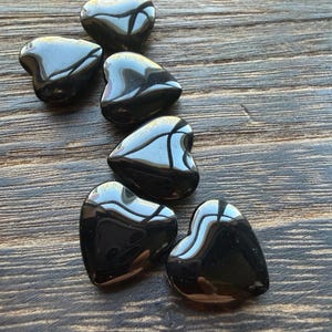 May include: Six glossy, black heart-shaped stones are arranged on a wooden surface. The stones reflect light, creating highlights and shadows. The hearts vary slightly in size and shape, suggesting they are natural or handcrafted.