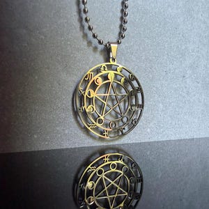 May include: A silver-toned metal pendant necklace featuring a circular design with a central pentagram surrounded by small circular moon phase icons and zodiac symbols along the outer edge, suspended from a matching silver-toned ball chain.