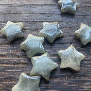 May include: A selection of small, star-shaped stones in a translucent, grey-green colour. The polished stones have a smooth, glossy surface and are arranged on a dark wooden surface. The stars vary slightly in size and shape.