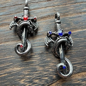 May include: Two silver-toned pendant necklaces featuring a sword design with a dragon wrapped around the blade. One pendant has red gemstones, the other has blue gemstones. The pendants are displayed on a dark wood surface.