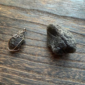 May include: Two dark gray tektite stones on a wooden surface. One is a raw, textured stone. The other is a pendant with a wire-wrapped design. The pendant has a teardrop-shaped tektite.