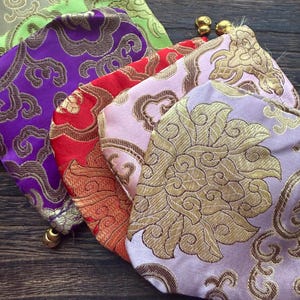 Puede incluir: Una colección de coloridas bolsas de tela con bordados dorados ornamentados. Las bolsas son en tonos morado, rojo, rosa, verde y naranja. Cada bolsa tiene una borla dorada y un cierre de cordón.